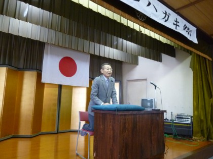 何と演台に座布団を敷き話す坂田道信さん